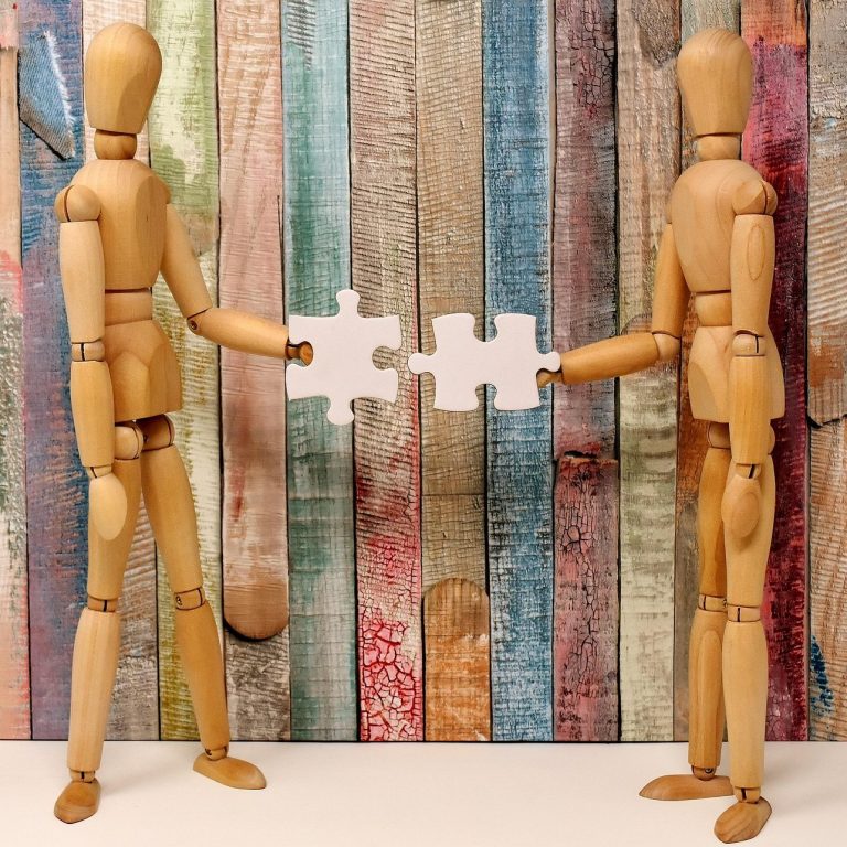 Two wooden mannequins exchange puzzle pieces against a colourful wooden backdrop.